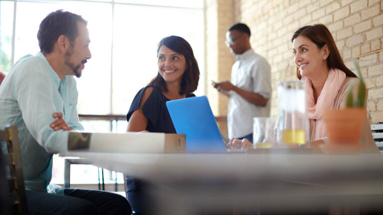 Three colleagues laughing and having a discussion while sitting around a laptop at a coffee shop table