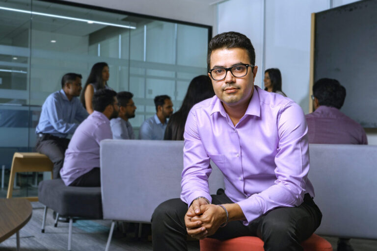 A male CSG employee sits in the Bangalore office as colleagues gather for discussion in the background.