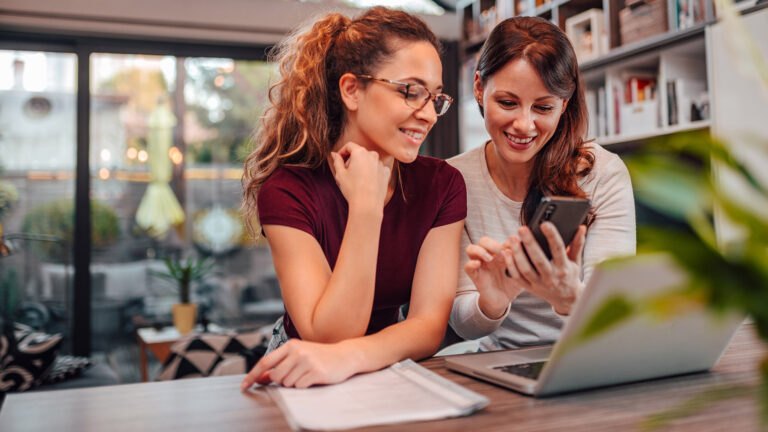 Two women share phone and laptop in coffee shop